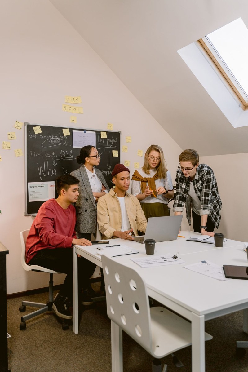 Five young professionals gathered around a laptop and sticky notes at a white table in a bright attic office