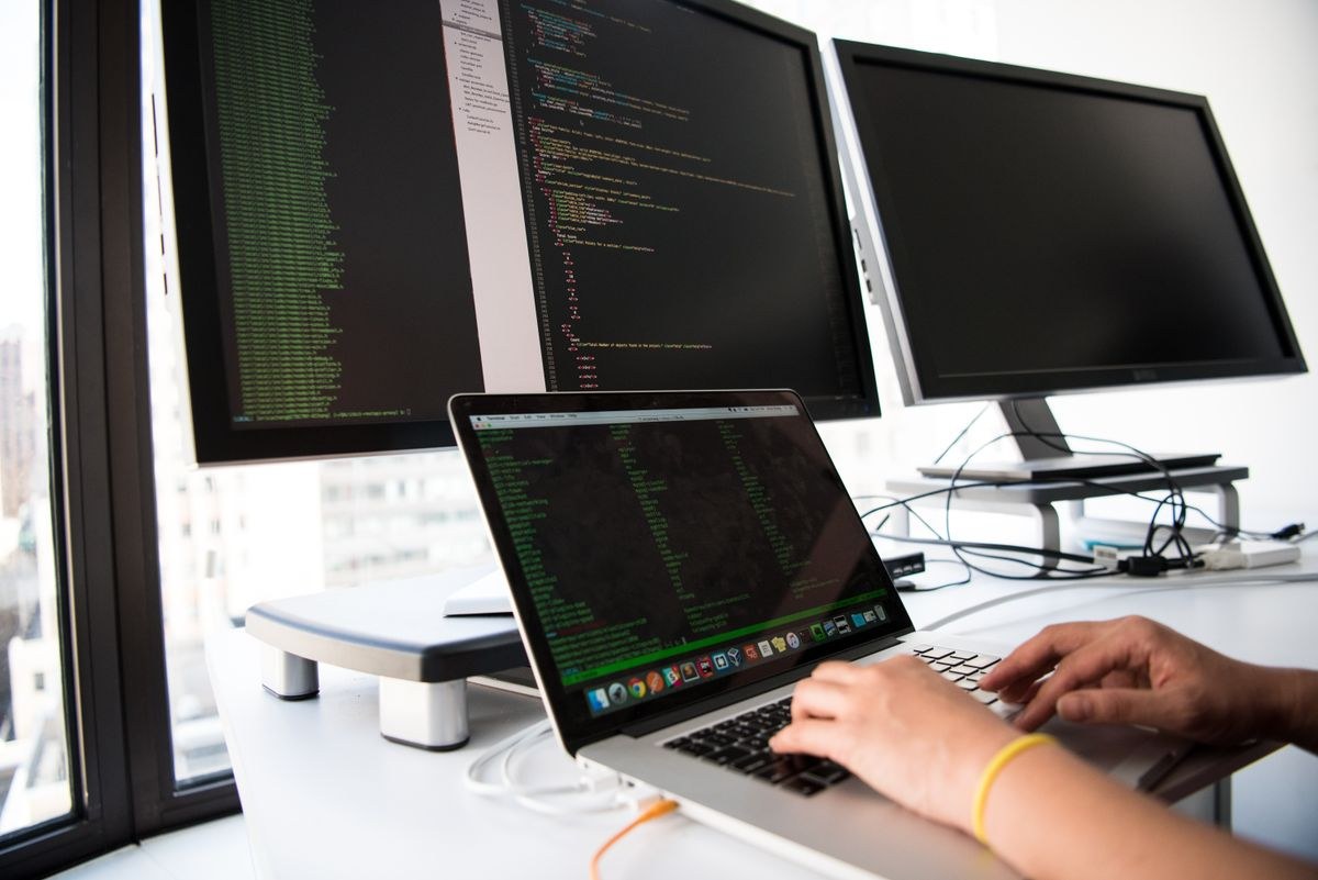 Person typing on a laptop at a desk with several monitors showing code