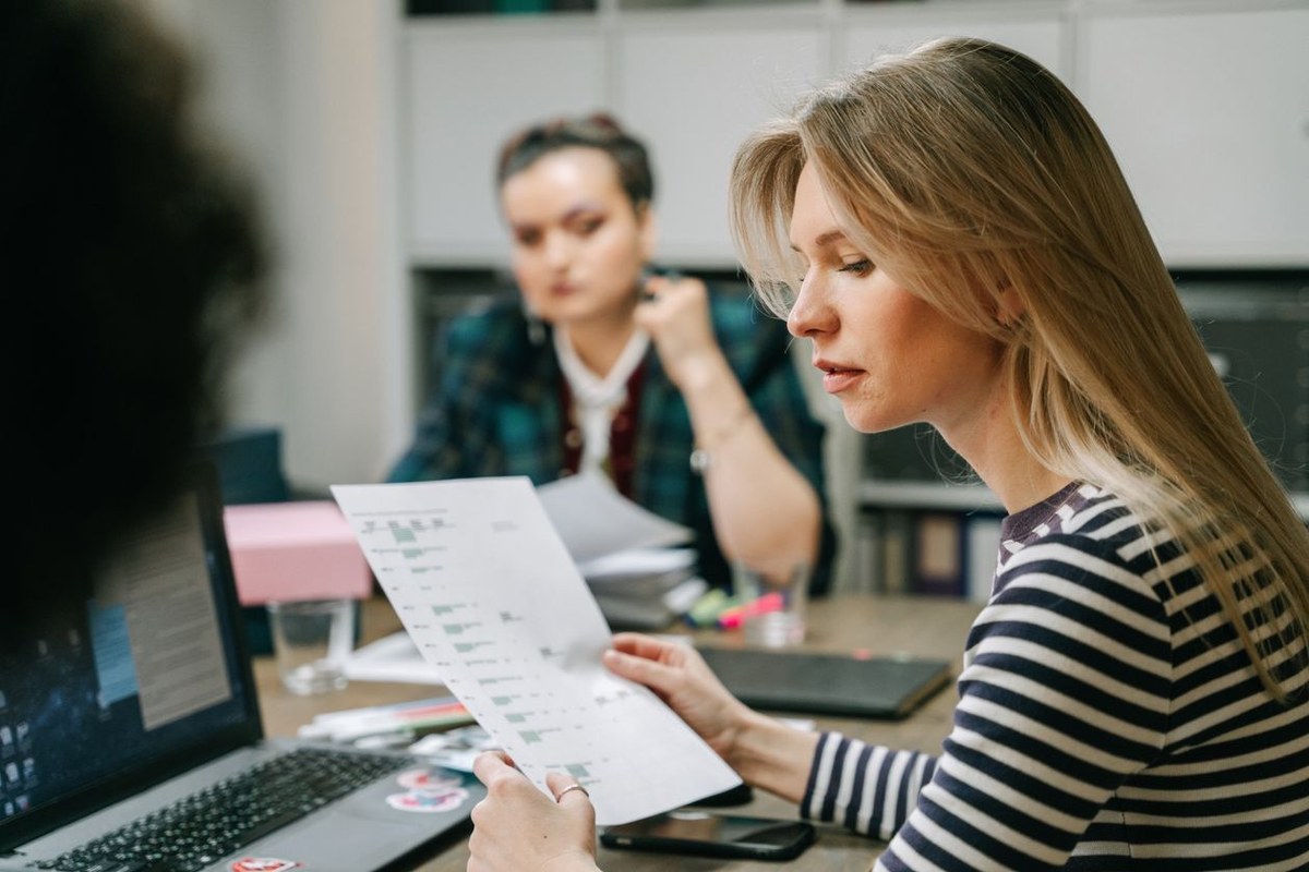 Woman in a striped shirt reviewing a printed business report with charts at a wooden desk beside an open laptop